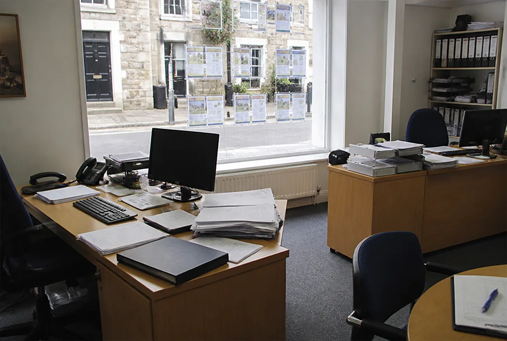 The inside of a small estate agency office with two desks, a window onto the street and paperwork on the side