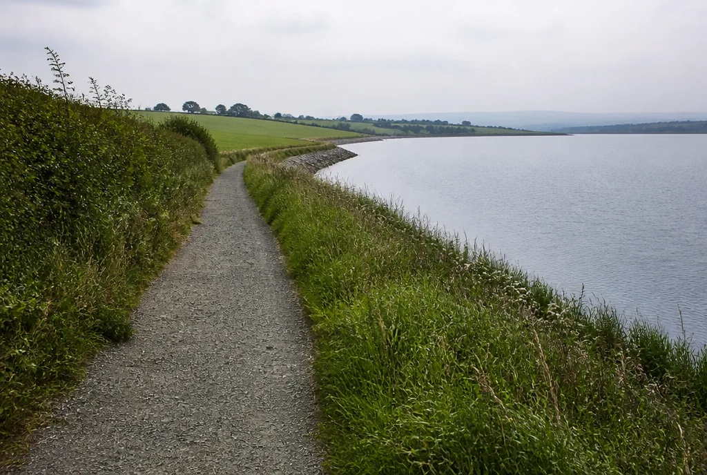 A footpath along the edge of a large reservoir with hedgerows and grazing fields beside it