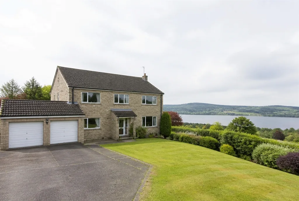 A detached stone-faced family house with a double garage and a lawn sloping towards distant open water