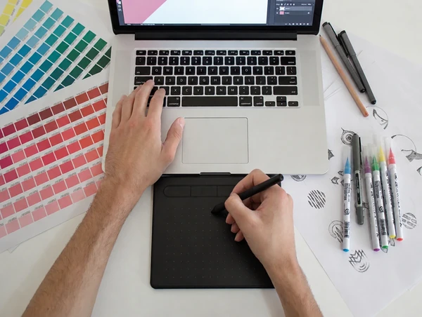 Hands draw with a pen on a graphics tablet beside a laptop, colour swatch strips and marker pens on the desk
