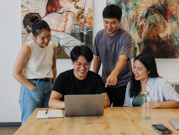 Four young Chinese Malaysian freelancers smile around a laptop at a light wooden table while a standing man points at the screen.