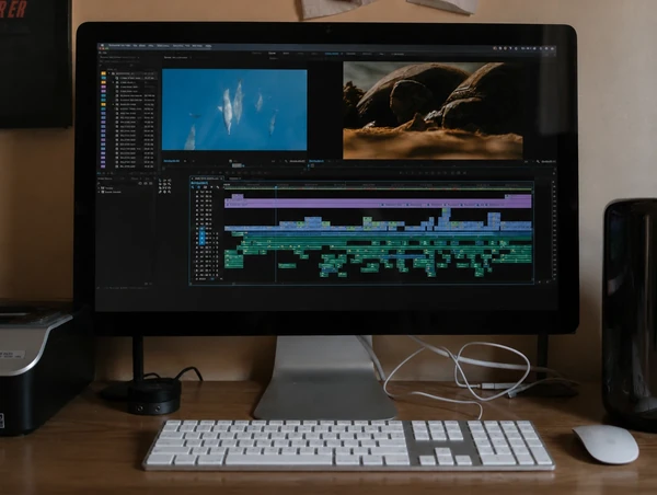 Video editing desk with a large monitor showing an edit timeline and preview clips, with a keyboard and mouse in front